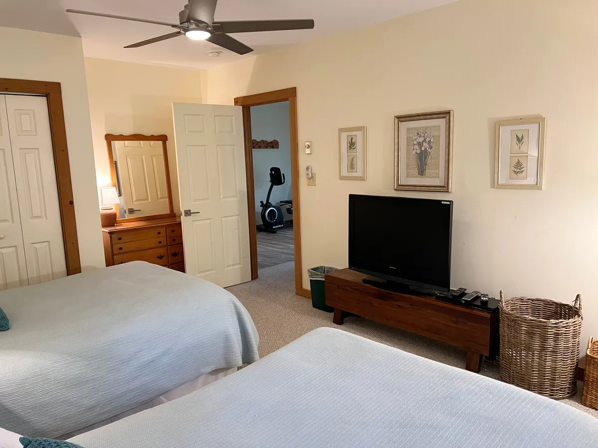 A welcoming room with two beds dressed in light blue covers, a wooden dresser, flat-screen TV, and decorative wall art. The arrangement is ideal for families or groups, with ample space for children, teens, or extra guests.