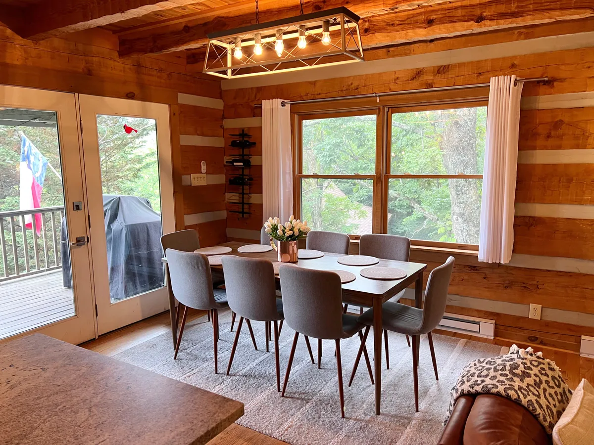 A sunlit dining area featuring a wooden table set beside large windows, allowing natural light to pour in and providing glimpses of the surrounding scenery. The space is dressed with a delicate floral centerpiece and a stylish light fixture, contributing to the warm, inviting ambiance ideal for intimate meals.