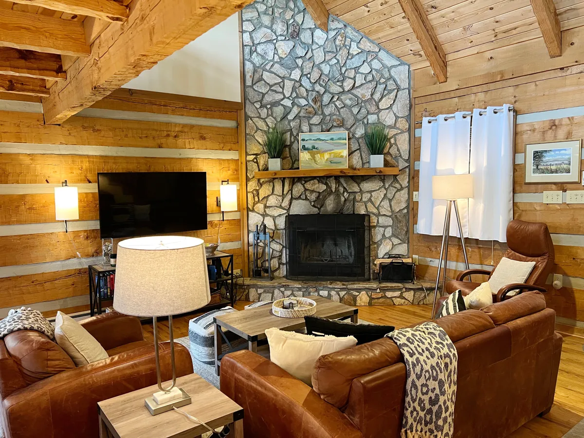 A warm, welcoming den centered around a striking stone fireplace, surrounded by comfortable leather sofas. A large TV is mounted above the mantle, with rustic wooden beams accenting the ceiling. The setting evokes evenings spent relaxing by the fire or enjoying a movie with the whole family.