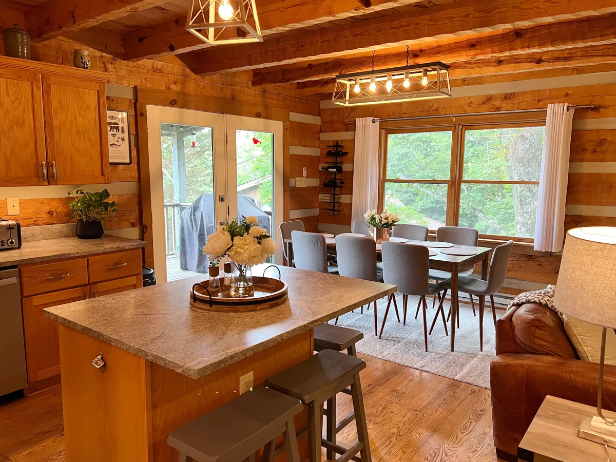 A light-filled dining and kitchen area featuring a generous island, a long dining table, wooden beams, and large windows looking out towards the natural surroundings. Decorative lighting and comfortable seating invite extended family meals and social gatherings.