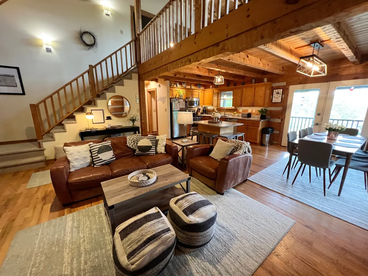 A wide-angle perspective of the open-plan living, dining, and kitchen area featuring leather sofas, a rustic wooden dining table, exposed beams, and a glimpse of mountain views through the windows.