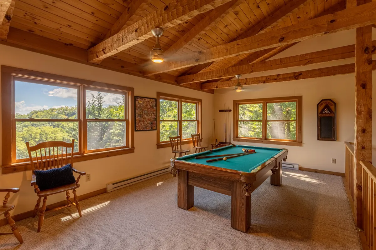 This image centers on a pool table set in a loft-like space, with large windows that showcase beautiful views and wooden ceiling beams that add rustic charm. The area looks welcoming, perfect for socializing or friendly competition, with natural light pouring in and the surrounding landscape visible.