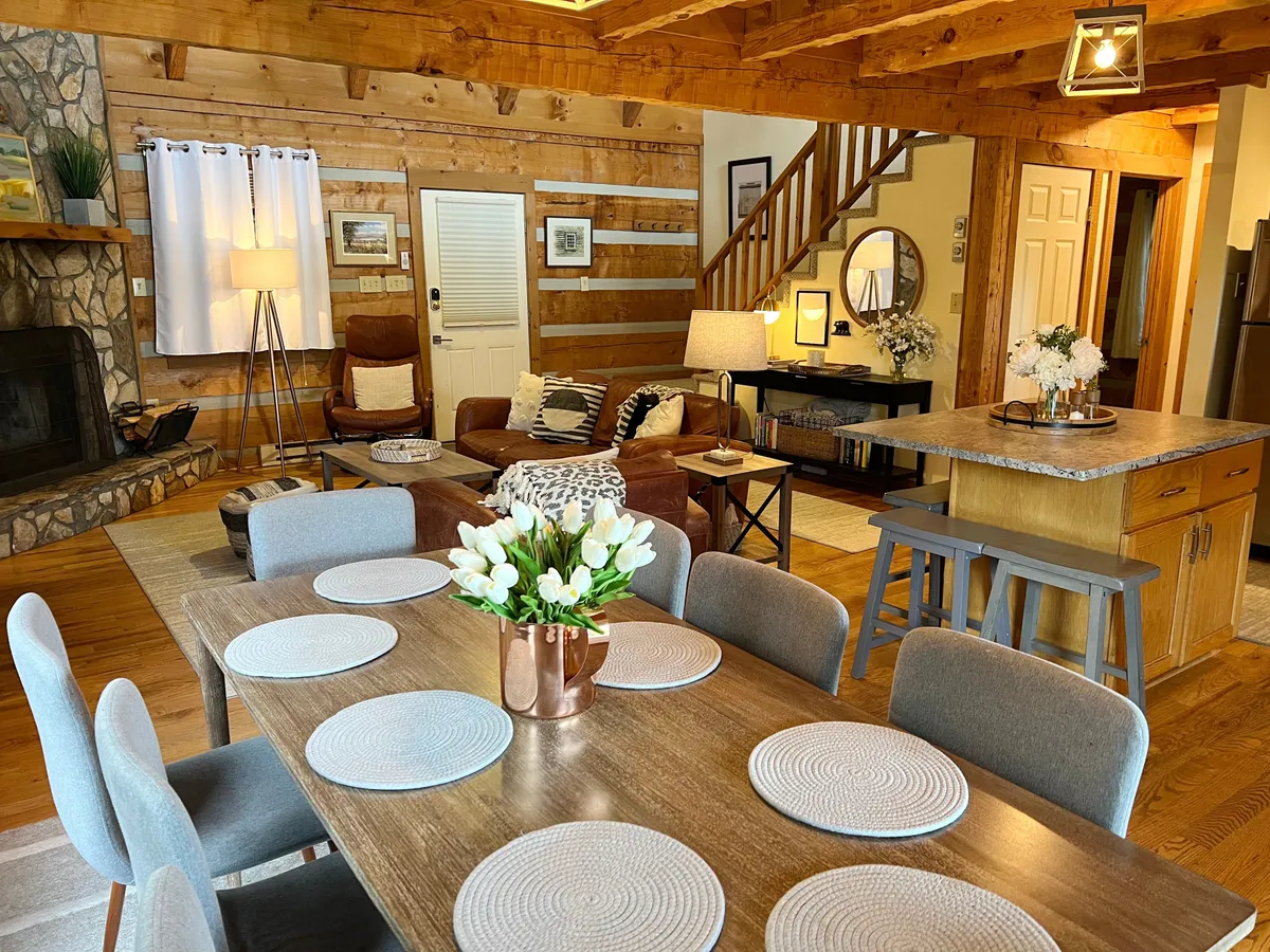 This inviting interior scene features a spacious dining table adorned with flowers, set next to a grand stone fireplace and a comfortable seating area. The wooden staircase hints at multiple levels, while abundant seating encourages group gatherings and shared meals. Soft lighting and warm tones create a cozy, communal atmosphere.