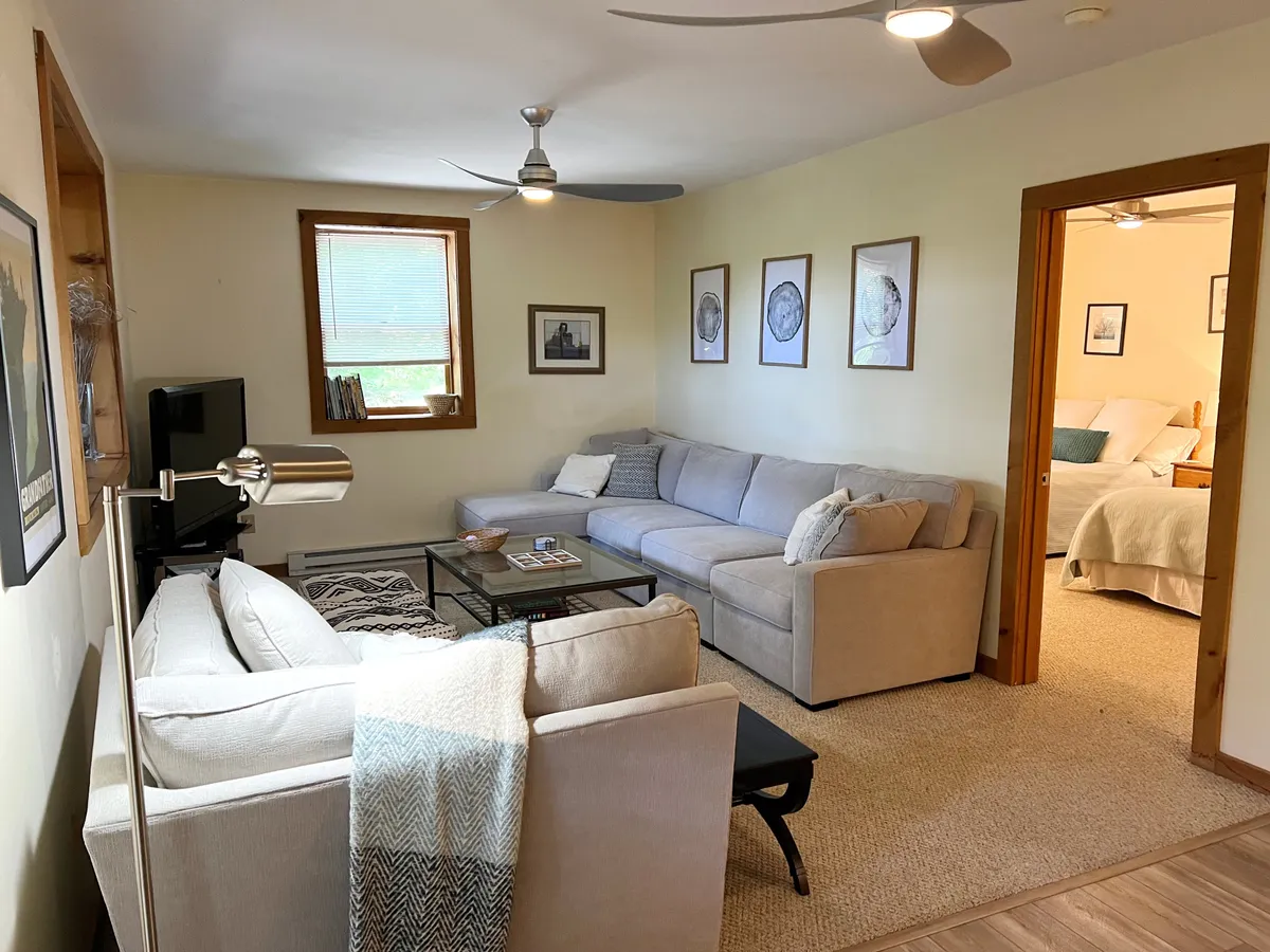 A comfortable den featuring a plush gray sectional sofa, a glass coffee table, tasteful wall art, and a window that lets in natural light. The setting feels intimate and perfect for movie nights, reading, or downtime, echoing the den's flexible, family-friendly purpose.