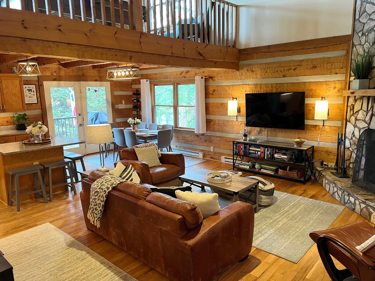 A panoramic view of an open-plan space featuring a large leather sofa facing a stone fireplace, with the adjacent dining area surrounded by bright windows. The space demonstrates seamless flow between the living, dining, and kitchen areas under high ceilings.