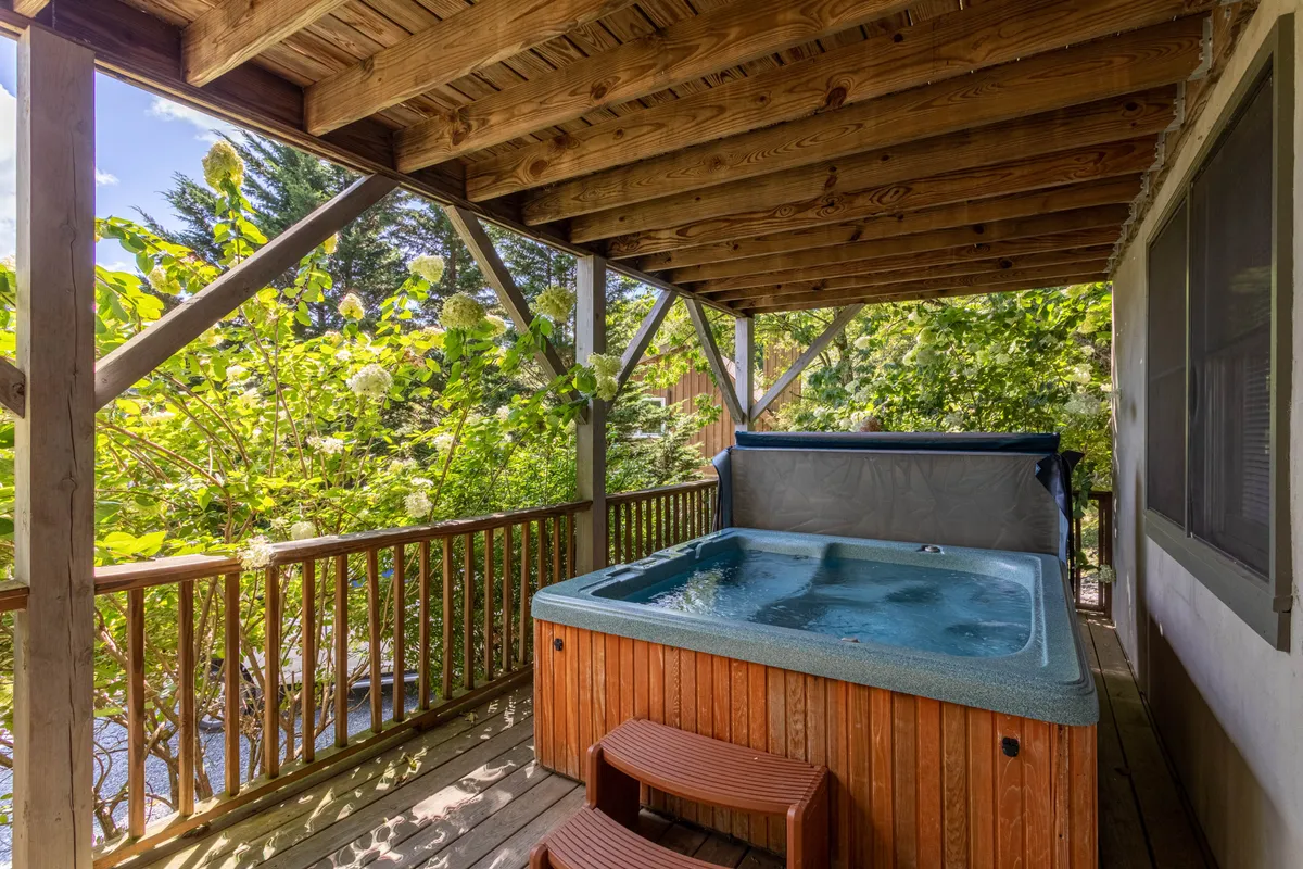 This exterior image features a wooden deck nestled in lush greenery, with a prominent hot tub as the focal point. The scene conveys relaxation and connection to nature, with the hot tub positioned for scenic views and inviting social interaction. The deck's spaciousness suggests ample room for outdoor gatherings, reinforcing the theme of al fresco mountain living.