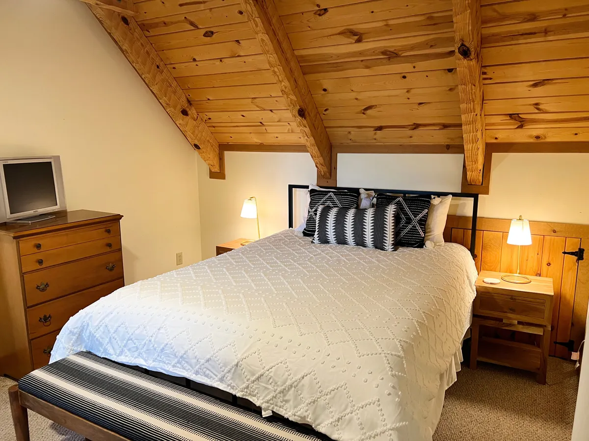 A welcoming bedroom with a plush white quilt, patterned pillows, and rustic wooden beams overhead. Soft morning light filters in, creating an atmosphere perfect for slow mornings in bed. Nightstands with lamps add to the cozy, restful feeling.