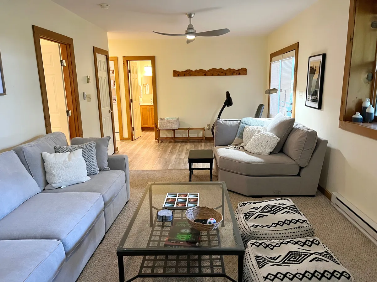 A cozy living area with a plush sofa, accent chairs, and a coffee table set under a stylish light fixture. The arrangement encourages relaxation, conversation, and social activities like board games or reading together. Natural light and inviting décor make it a perfect hub for group interaction.