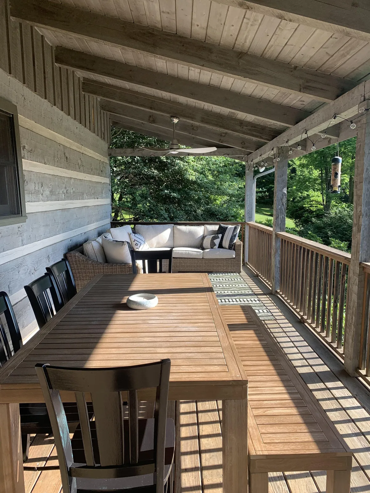 An expansive covered deck furnished with a wooden dining table, comfortable outdoor sofa, and surrounded by greenery, offering space for outdoor gatherings, meals, and relaxation while enjoying nature.