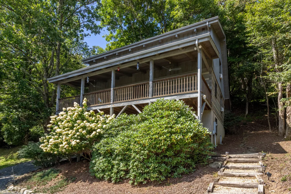 A wide wooden balcony overlooks mountain flora, with a flowering shrub adding vibrant color and a stone pathway winding below. The scene captures moments of relaxation, such as sipping coffee on the porch or enjoying conversation as the sun sets. The covered deck area is designed for comfort, supporting both quiet solitude and lively social gatherings.