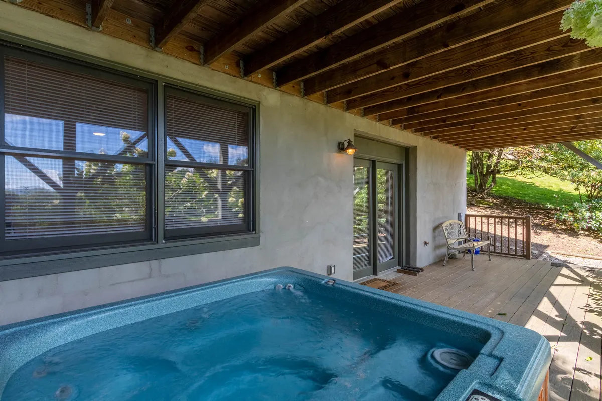 A serene view of the cabin's wooden deck, complete with a bubbling hot tub and seating area. The windows with blinds reflect the peaceful outdoor environment, perfect for unwinding after a day of exploration.