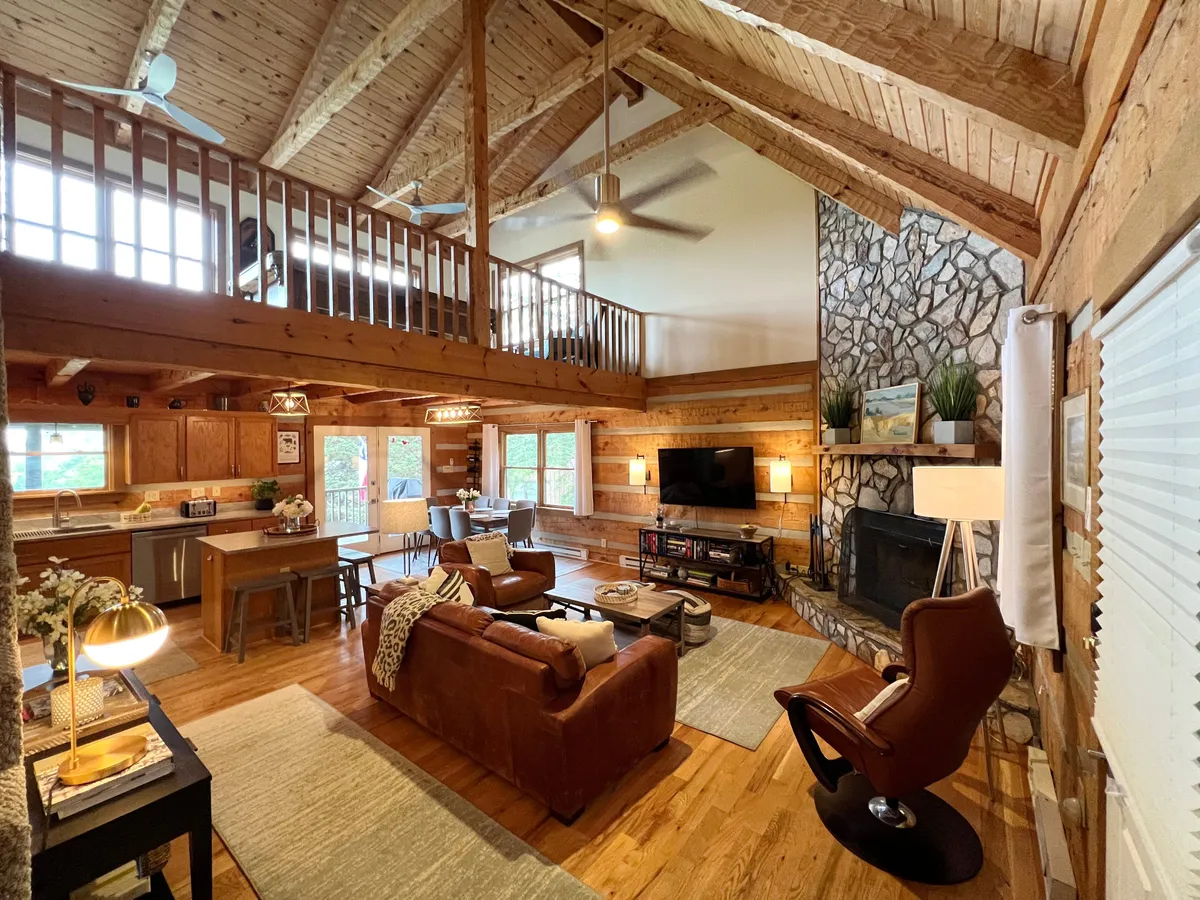 A spacious living room with a large stone wood-burning fireplace, open loft area above, and a comfortable seating area of plush sofas. Sunlight pours in from windows, highlighting the high vaulted ceilings and creating a warm, inviting atmosphere ideal for gatherings.