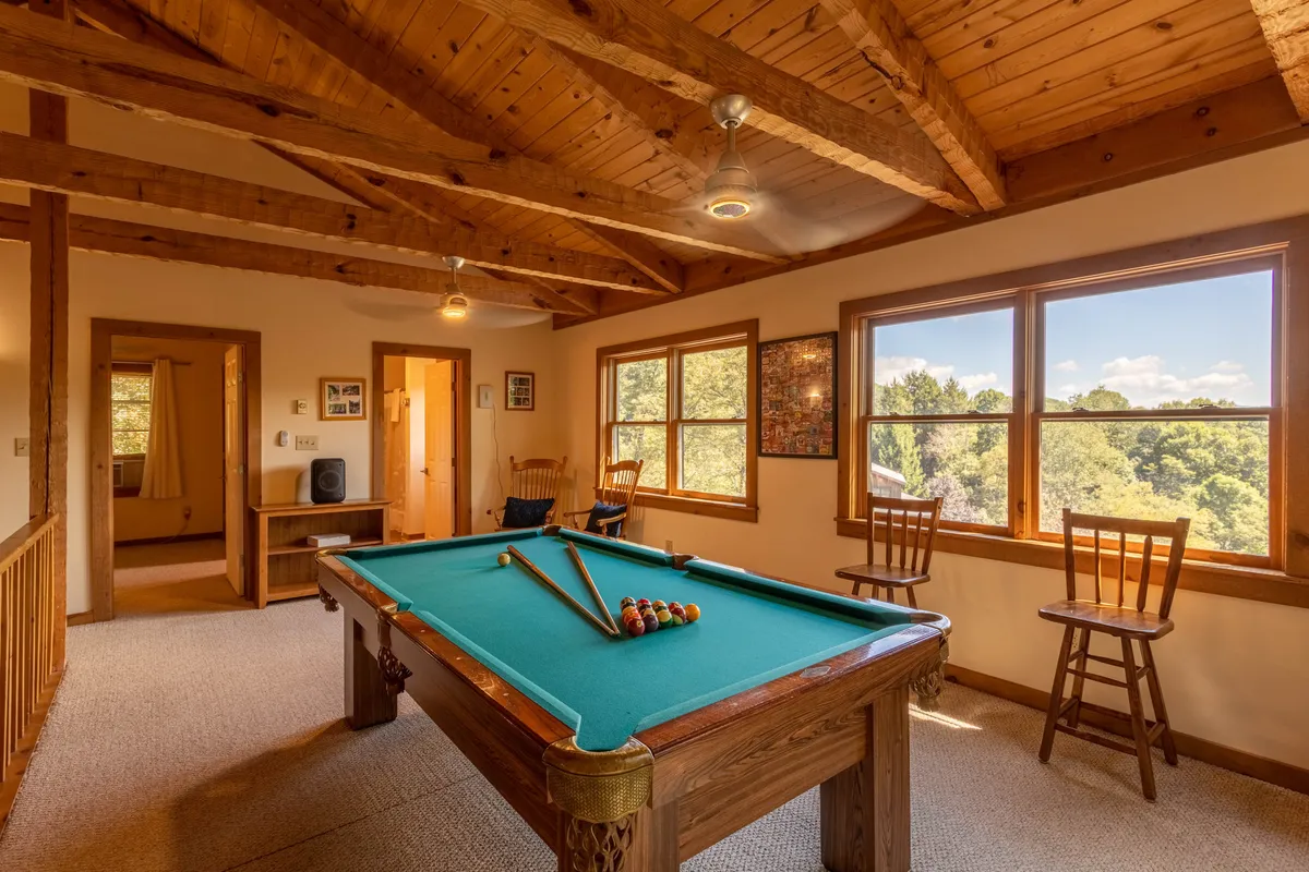 A relaxed loft area with a pool table, bar stools, large windows, and exposed wooden beams. The setup suggests a casual atmosphere ideal for late-night games, reading, or enjoying the scenery. Abundant light and seating options make it versatile for group gatherings or quiet time.